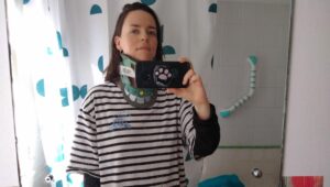 A mirror selfie of Mika in the bathroom. Mika is wearing a blue and white striped t-shirt and a neck brace. Mika has shoulder-length hair and smiles while holding the smartphone in front of their body with a pink paw print on it.